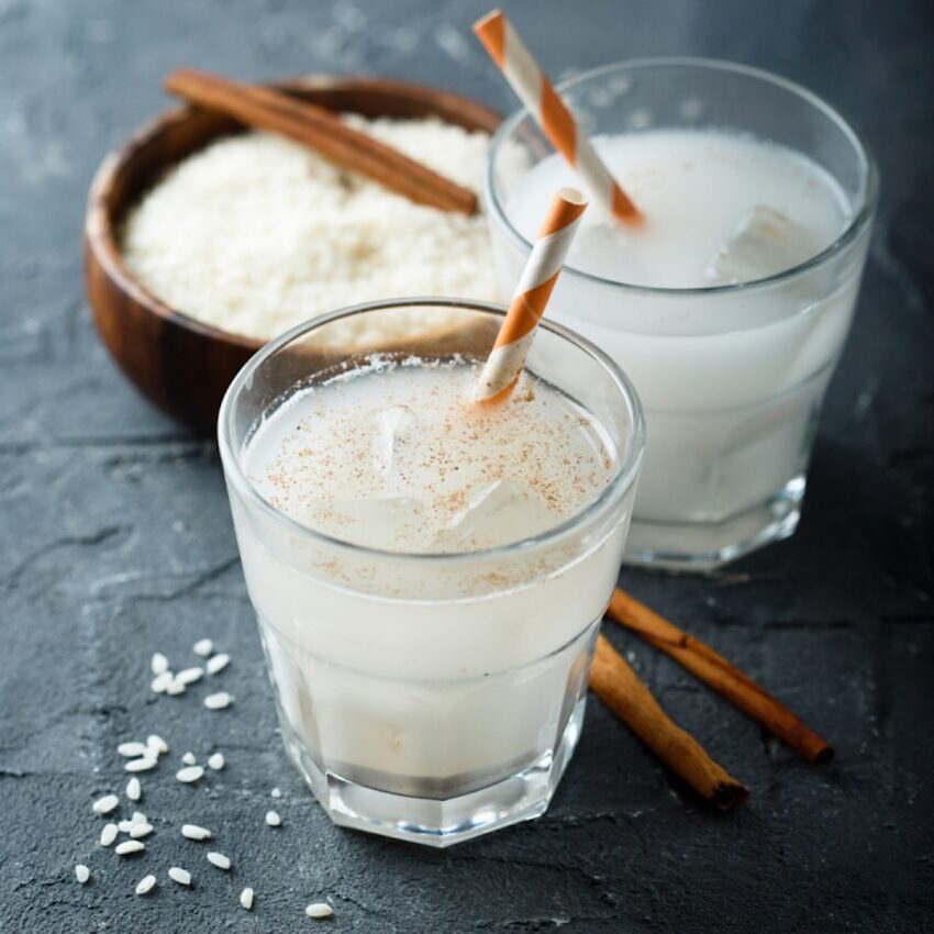 Two glasses of horchata with cinnamon stick stirrers and growl of rice milk in background.