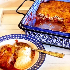 Bowl of strawberry rhubarb cobbler with ice cream and the baking dish of cobbler behind it.