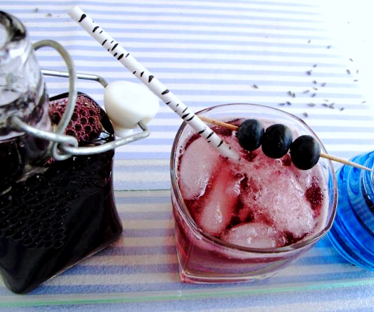 Glass of sparkling water with ice cubes and blueberry syrup and a bottle of blueberry syrup next to it.