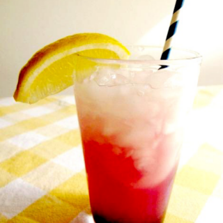 Glass of lemonade with blueberry syrup garnished with wedge of lemon and a striped straw.