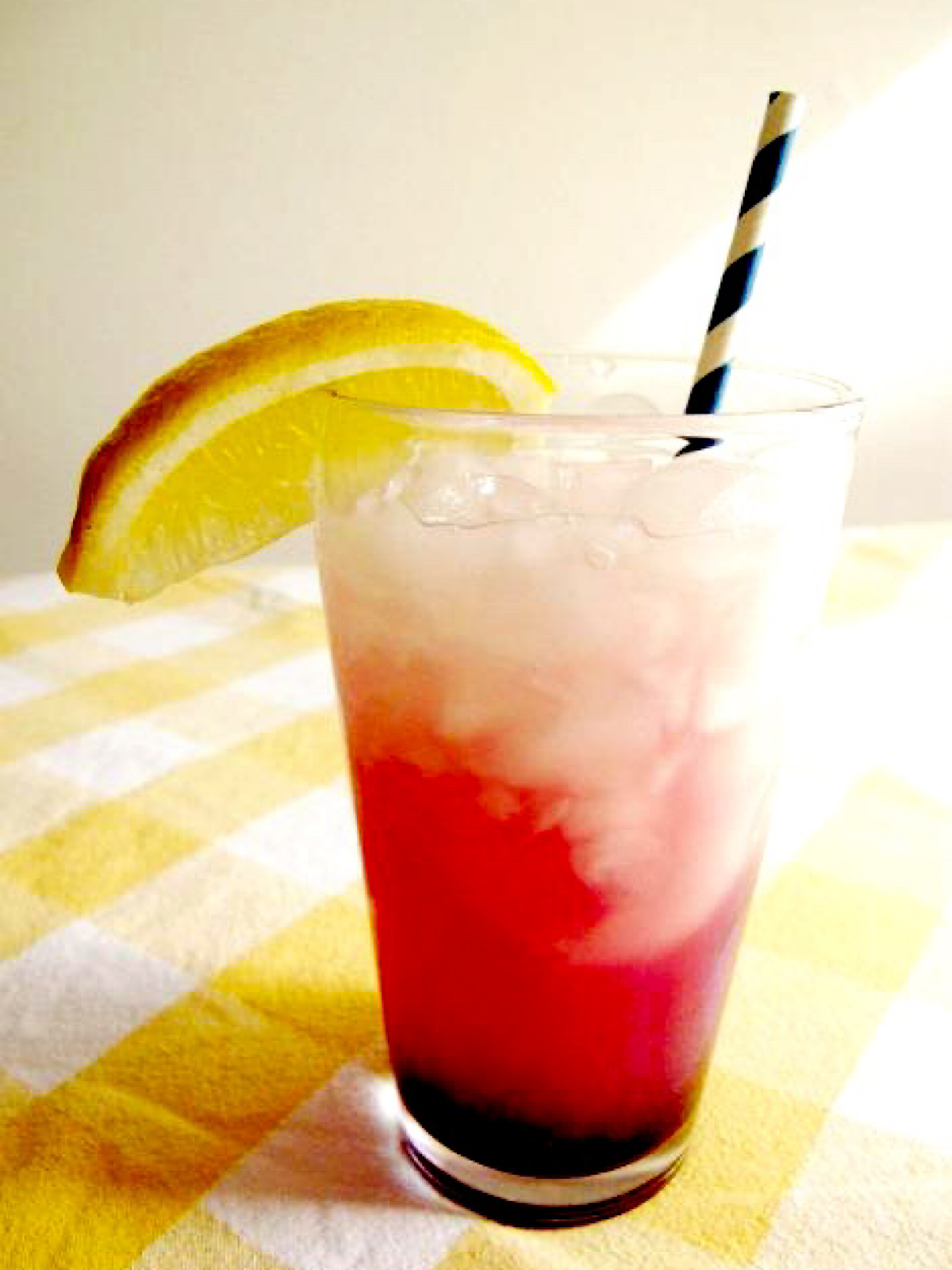 Glass of lemonade with blueberry syrup garnished with wedge of lemon and a striped straw.