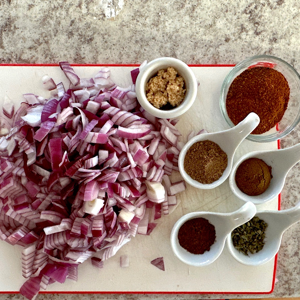 Chopped onions, minced garlic and a range of spices to use in chile, on a white cutting board.
