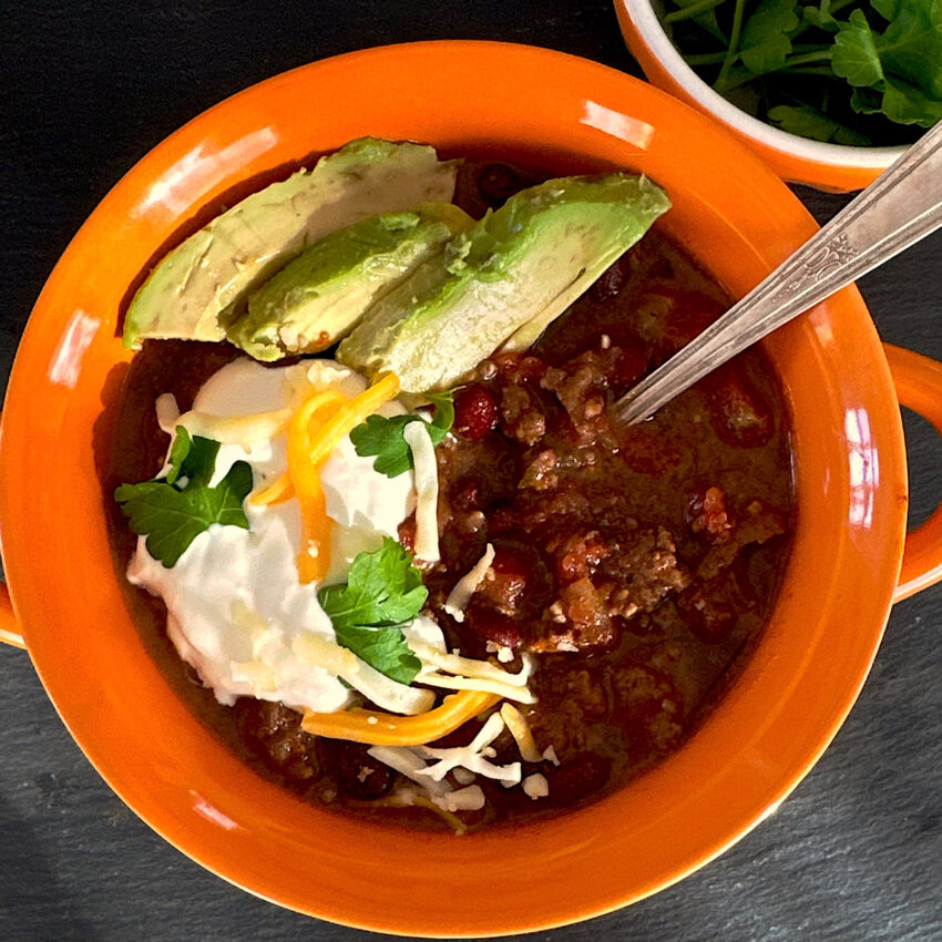 Orange bowl of chile garnished with sour cream, cilantro, cheese and avocados.