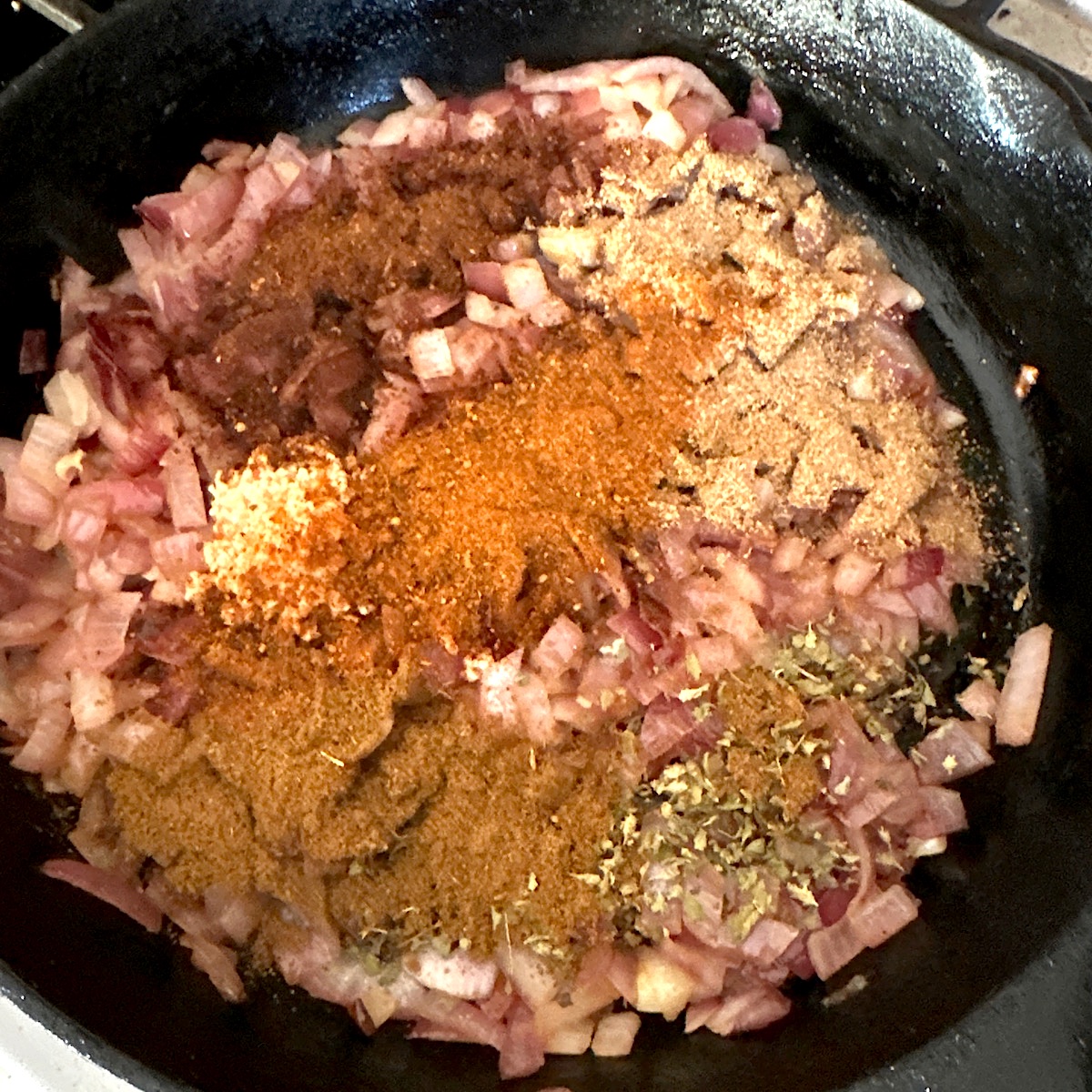 A range of chile spices added to chopped onions in a black cast iron skillet.