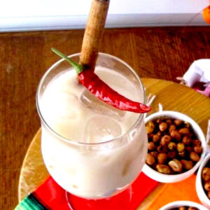 Tall glass of horchata with cinnamon stick and red pepper garnish and chile. Lime spiced peanuts in background.