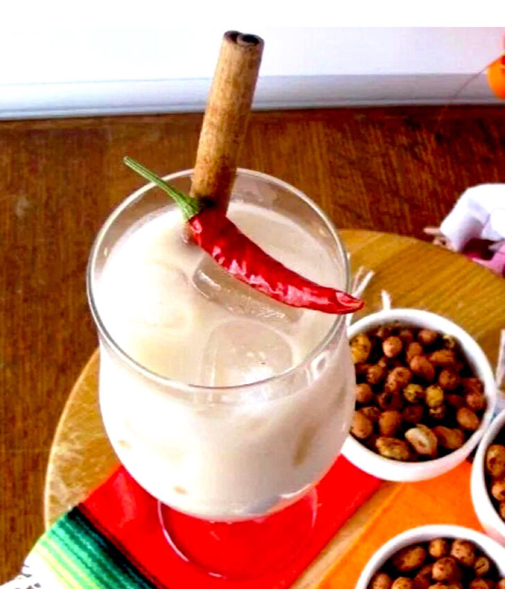Tall glass of horchata with cinnamon stick and red pepper garnish and chile lime peanuts in background.