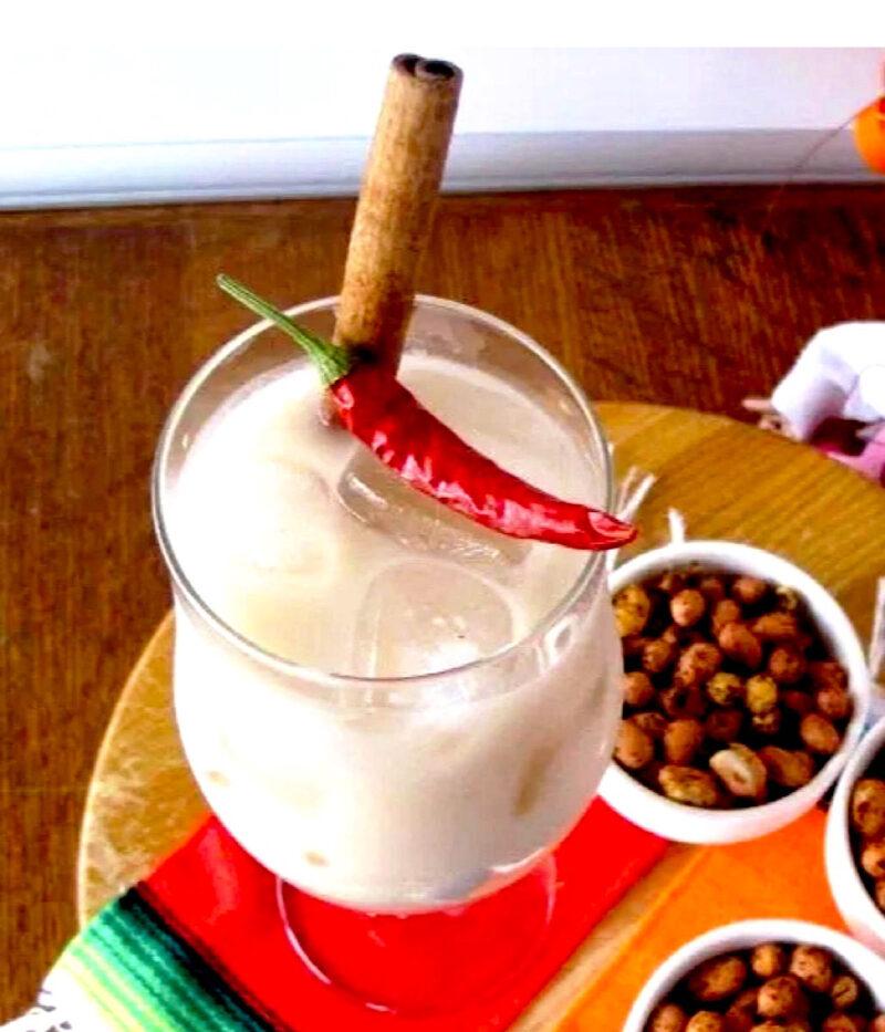 Tall glass of horchata with cinnamon stick and red pepper garnish and chile. Lime spiced peanuts in background.