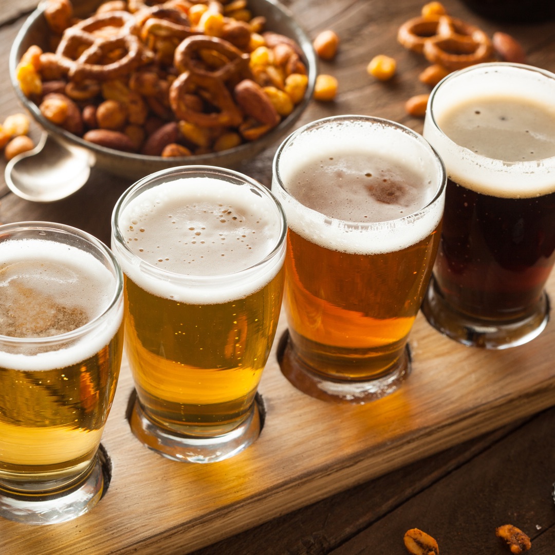 A flight of various beers with a bowl of pretzels in the background.