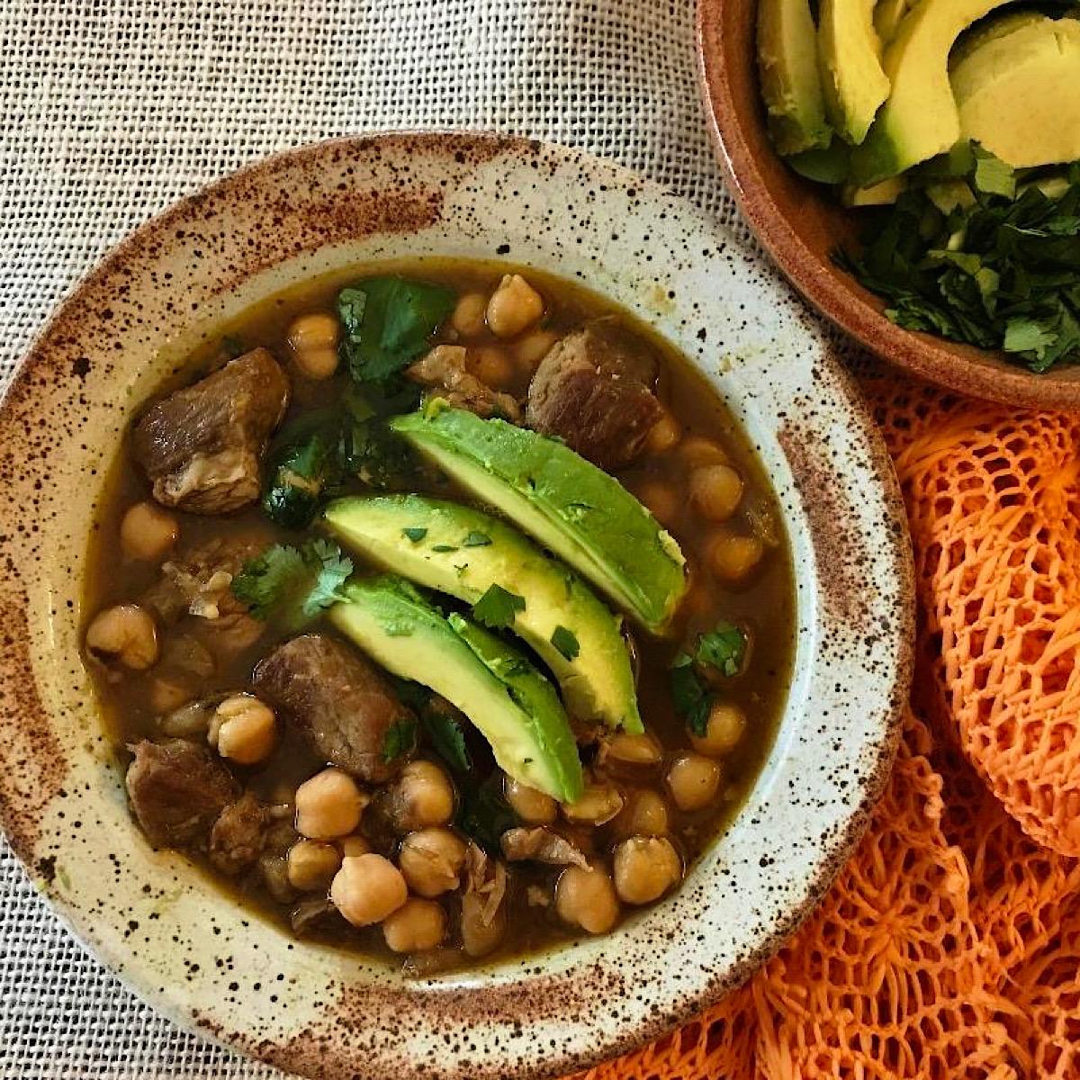 Bowl of pork posole with garbanzo beans and sliced avocado garnish