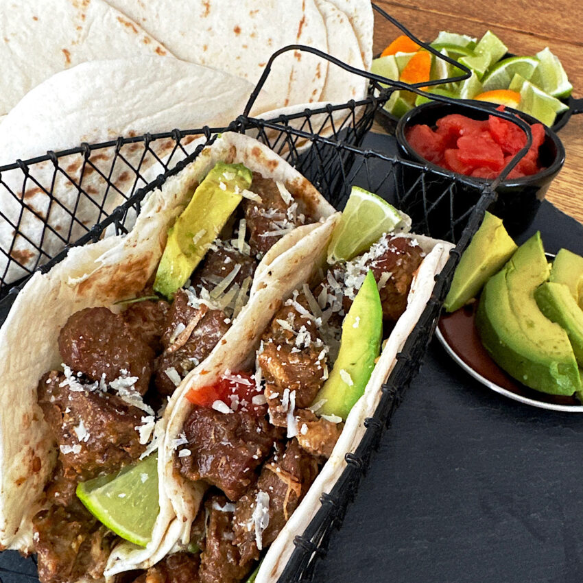 Two tacos in a black basket made with carnitas with avocados and tomatoes and tortillas in the background.