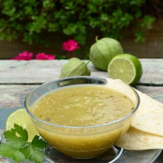 Glass bowl of chile verde sauce with tomatillos, lime and tortillas to the side.