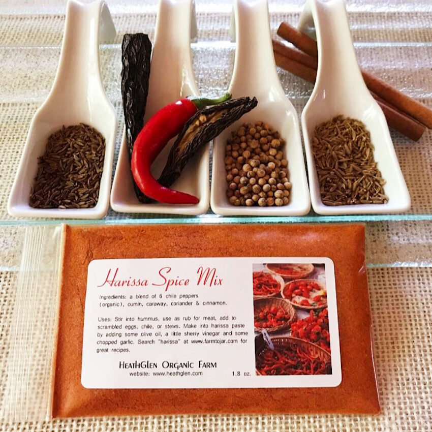 Harissa spice blend in a plastic bag with cumin, chile powder, coriander, and caraway seeds in white spoons.