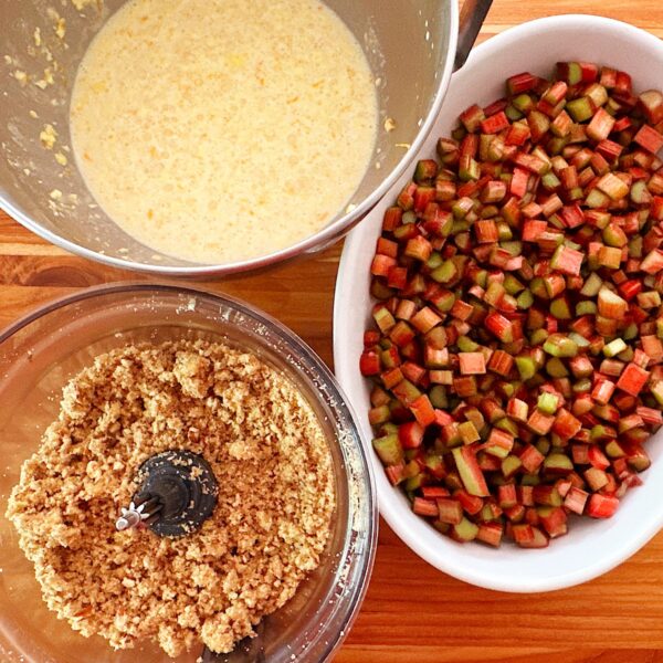 Baking dish with cut up rhubarb, food processor bowl of crumble topping, and bowl of custard mixture.