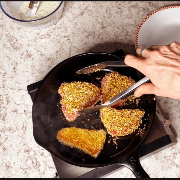 4 filets of nut-crusted tuna in a skillet with a hand using tongs to turn.
