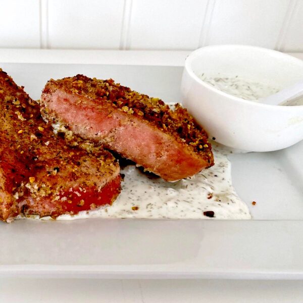 2 pistachio-crusted tuna steaks with a cup of sour cream dip on the side, all on a white plate.