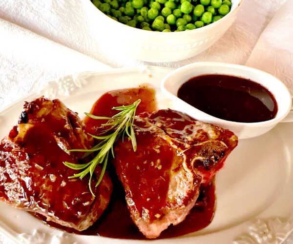 2 lamb chops drizzled with Cumberland sauce and a sprig of rosemary with a side of sauce and a bowl  of green peas in background.