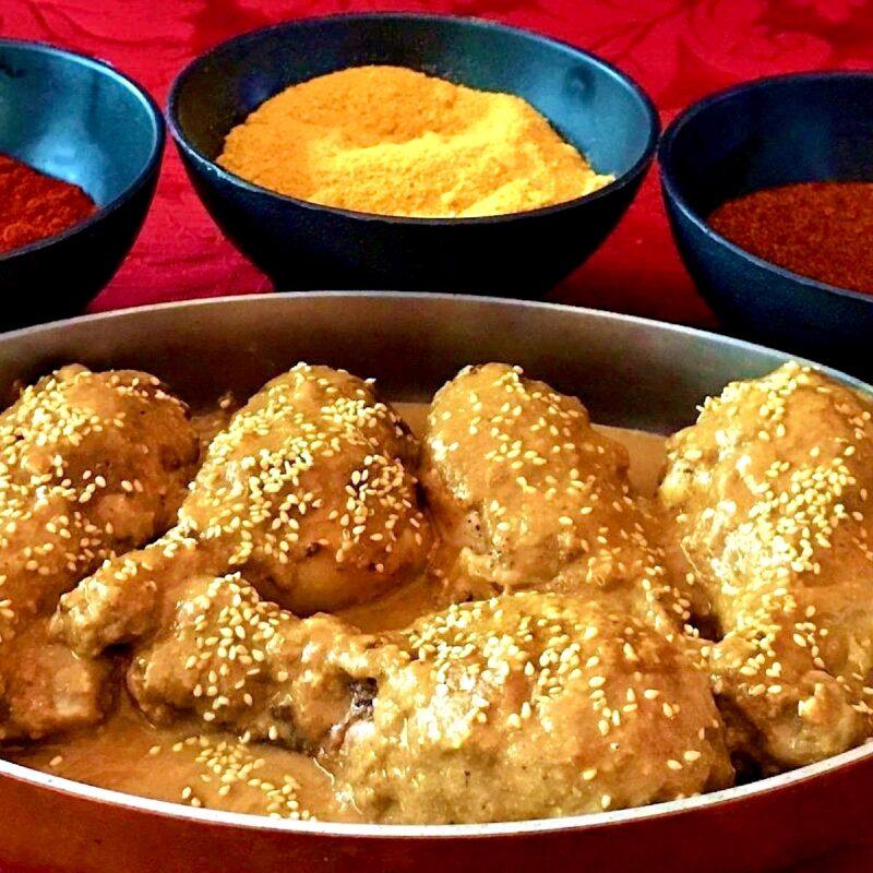 Chicken legs in a mole rojo sauce with spice blends in bowls behind the chicken.