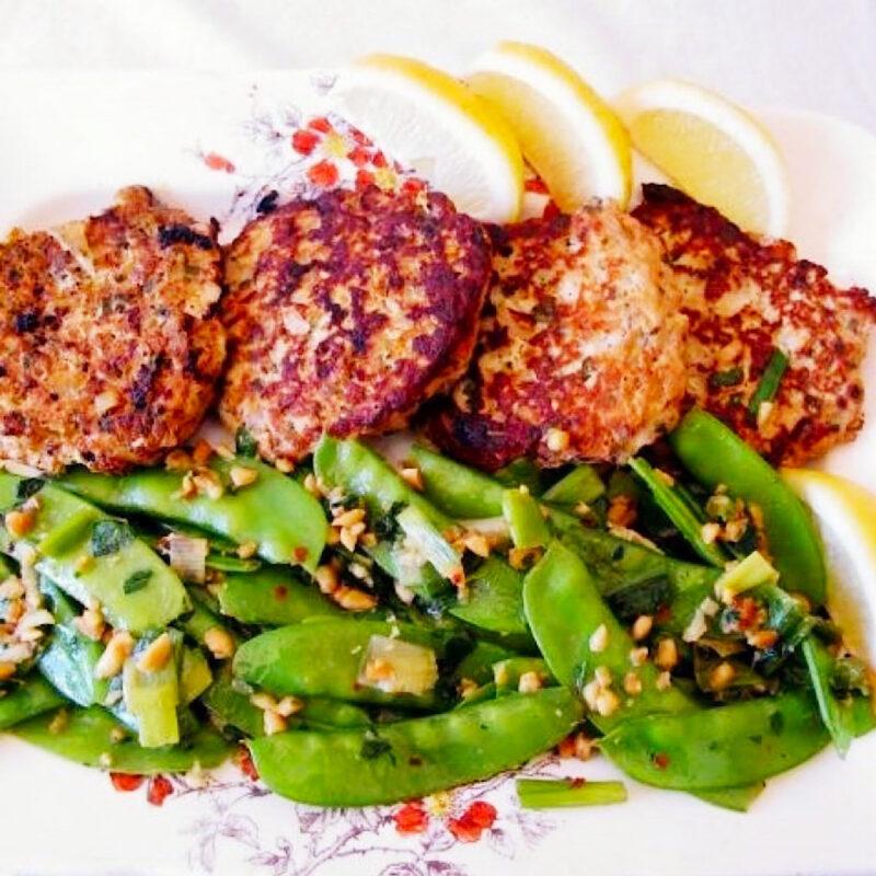 4 chcken patties with snow pea saute in front and lemon wedges behind.