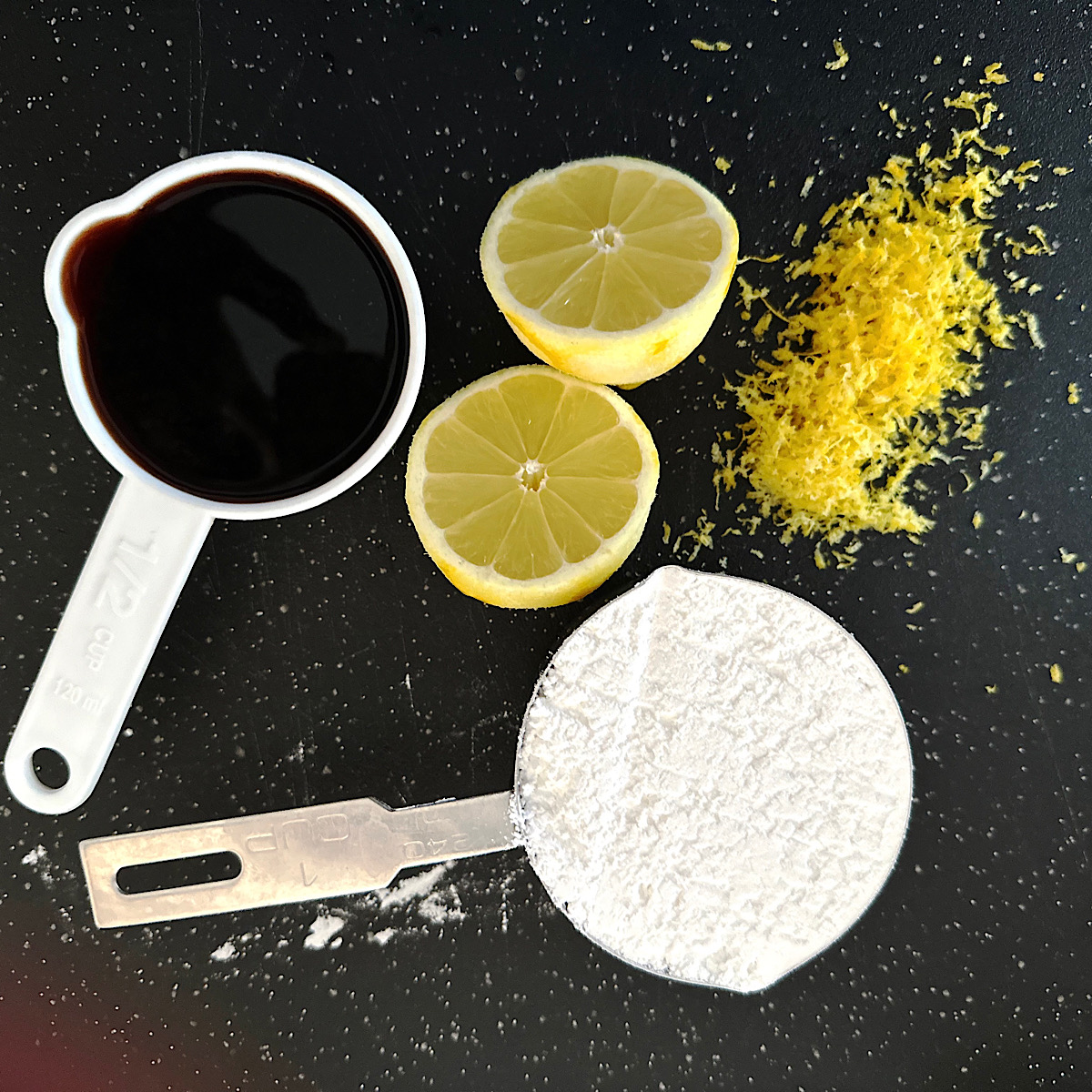 Balsamic vinegar, lemon and lemon zest, powdered sugar on a black surface.