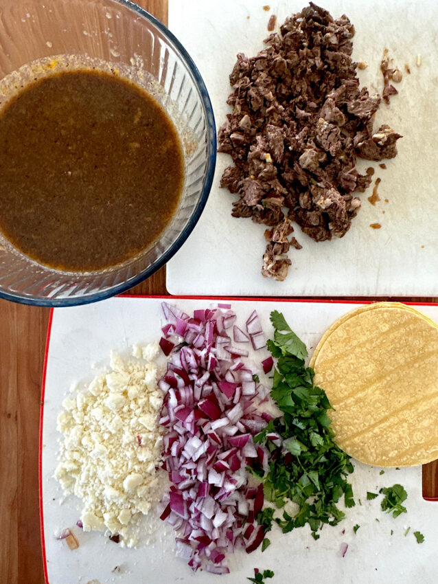 Ingredients for making birria quesatacos, including cheese, onions, cilantro, tortillas, meat, consommé.