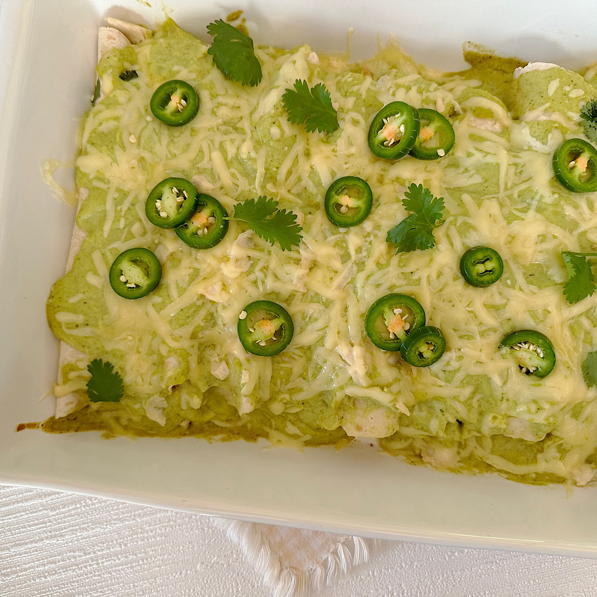 White baking dish of chicken enchiladas with a tomatillo sauce and topped with cheese.