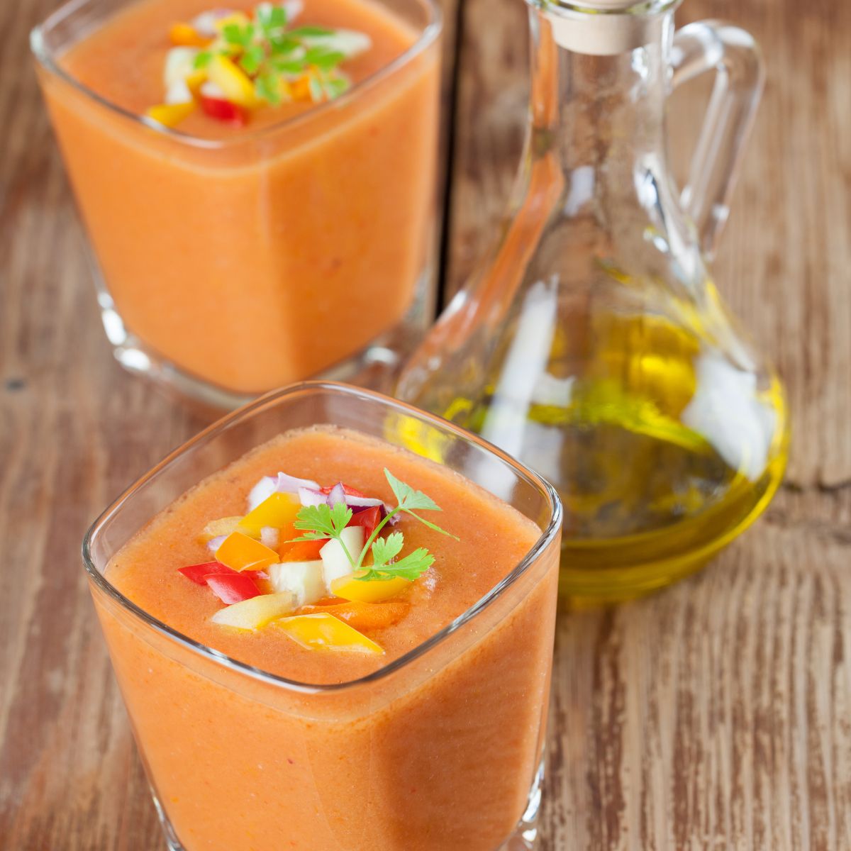 Two small glasses of gazpacho shooters with a carafe of olive oil on the side.