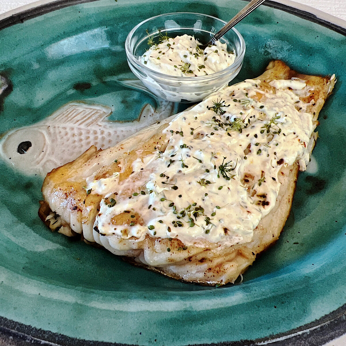 Filet of red snapper with sour cream dill sauce on a blue plate with a side of sauce in a small glass bowl.