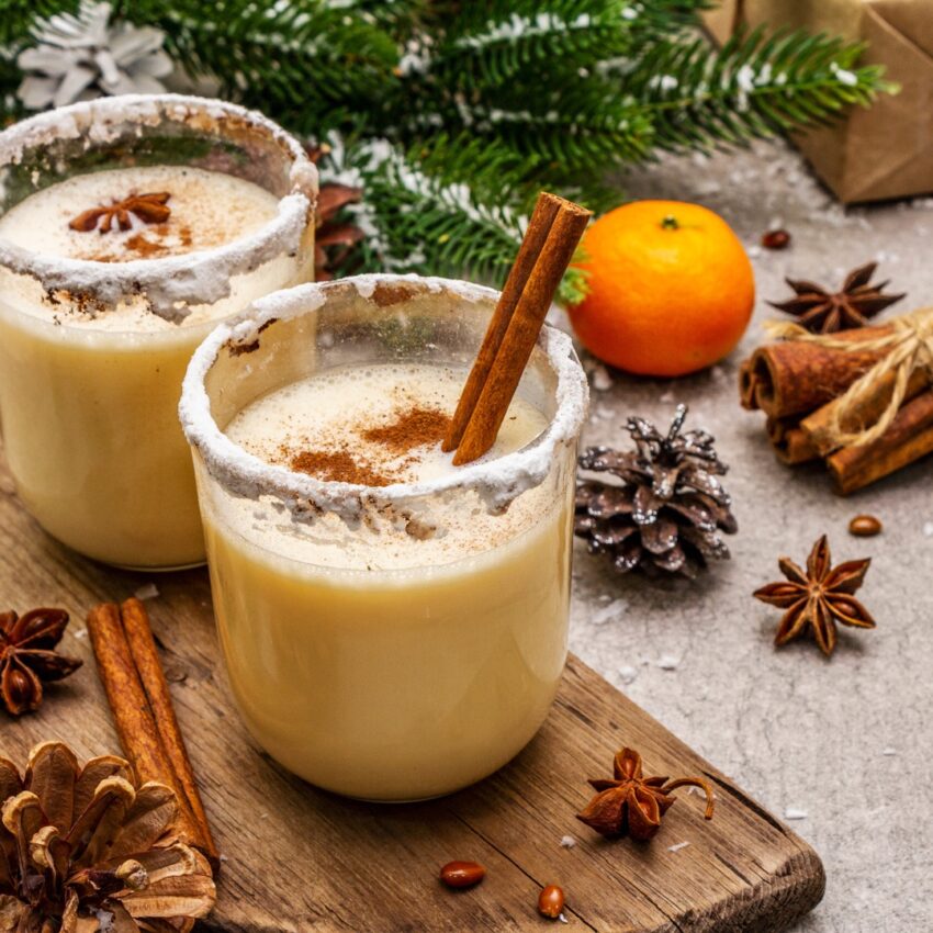 Two glasses of Dominican eggnog with cinnamon sticks, orange and spruce branches in background.