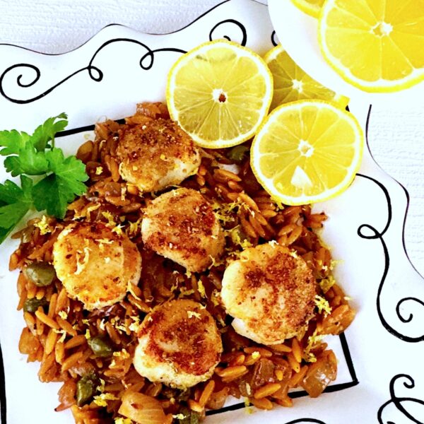 Pan seared scallops on a bed of lemon orzo on a white plate with black squiggles.