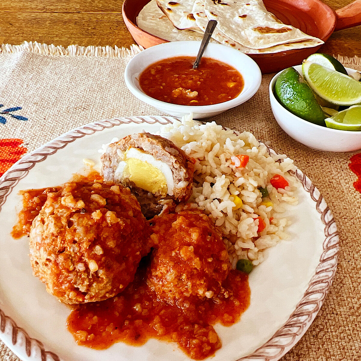 Two meatballs with red sauce and vegetable rice on the side with limes, sauce and tortillas in the background