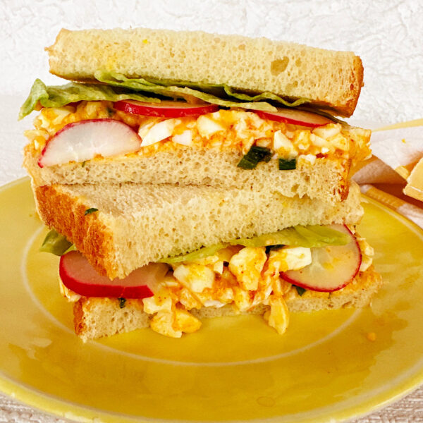 Two halves stacked on top of each other of egg salad sandiwich with radishes and scallions; on a yellow plate.