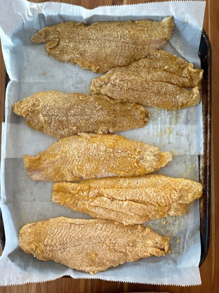 6 breaded catfish filets laid out on parchment paper on a cookie sheet.