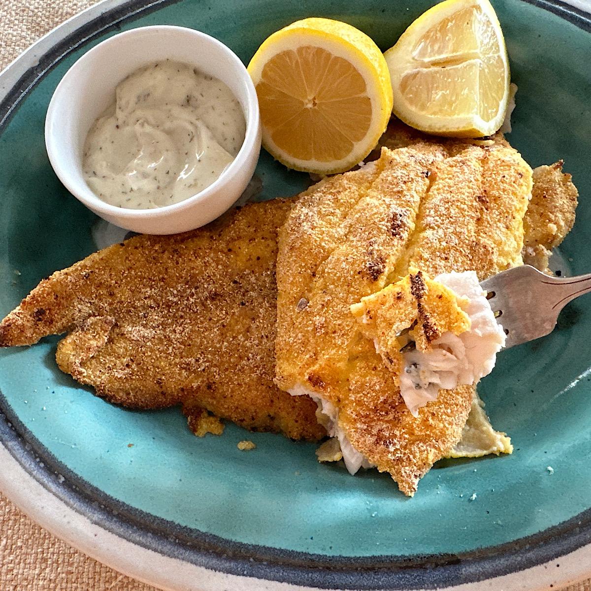 Two catfish filets breaded with a cornmeal-Mexican spice rub and baked, with a sour cream dip and 2 lemons on side on a blue plate.