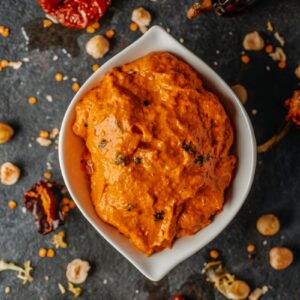 Romesco in white bowl surrounded by peppers and nuts.