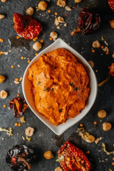 Romesco in white bowl surrounded by peppers and nuts.