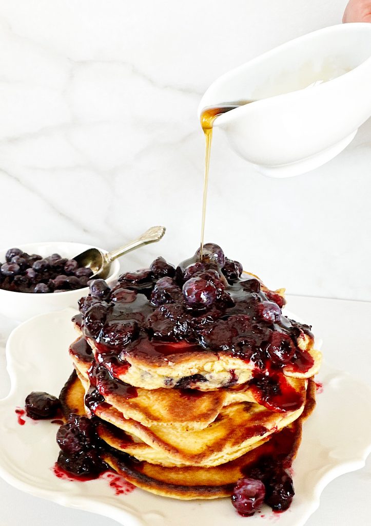 Stack of pancakes  with blueberry topping and syrup being poured over.