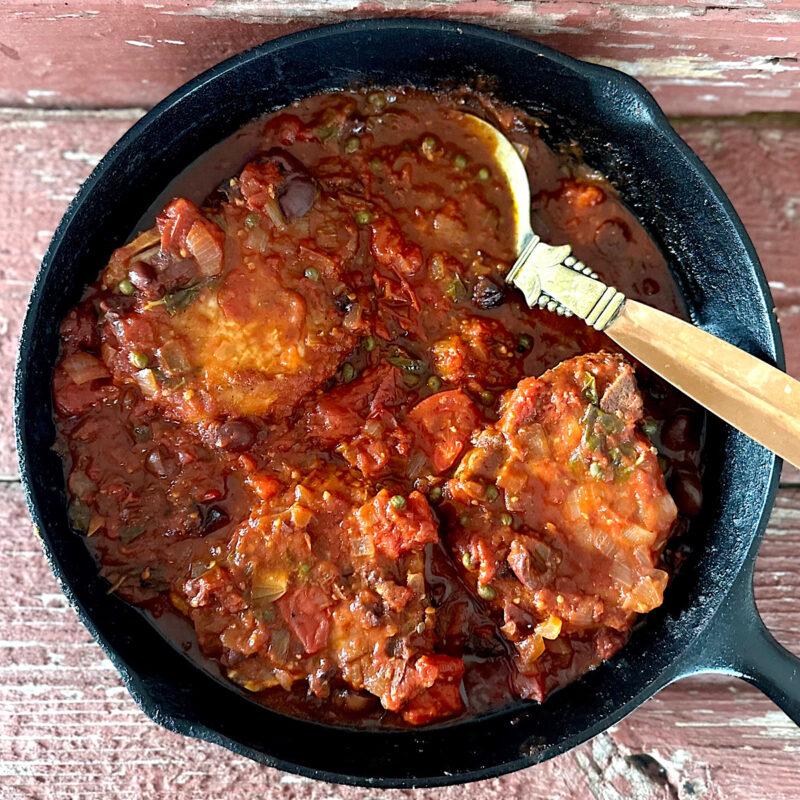 Pork chops in a skillet smothered with puttanesca sauce.