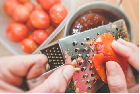 Close up photo of femail hand grating a tomato with a box grater.