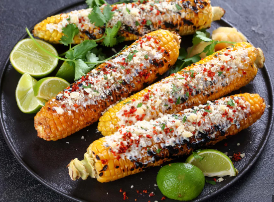 Photo of 4 Mexican grilled corn on cobs (called "elote") on a dark plate with sliced limes as a garnish.