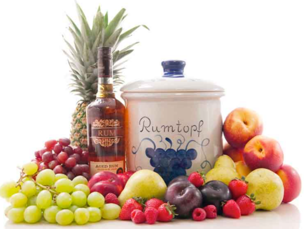 Photo of a rumtopf white ceramic pot surrounded by fresh fruit such as grapes, pears, apples, berries, pineapple.