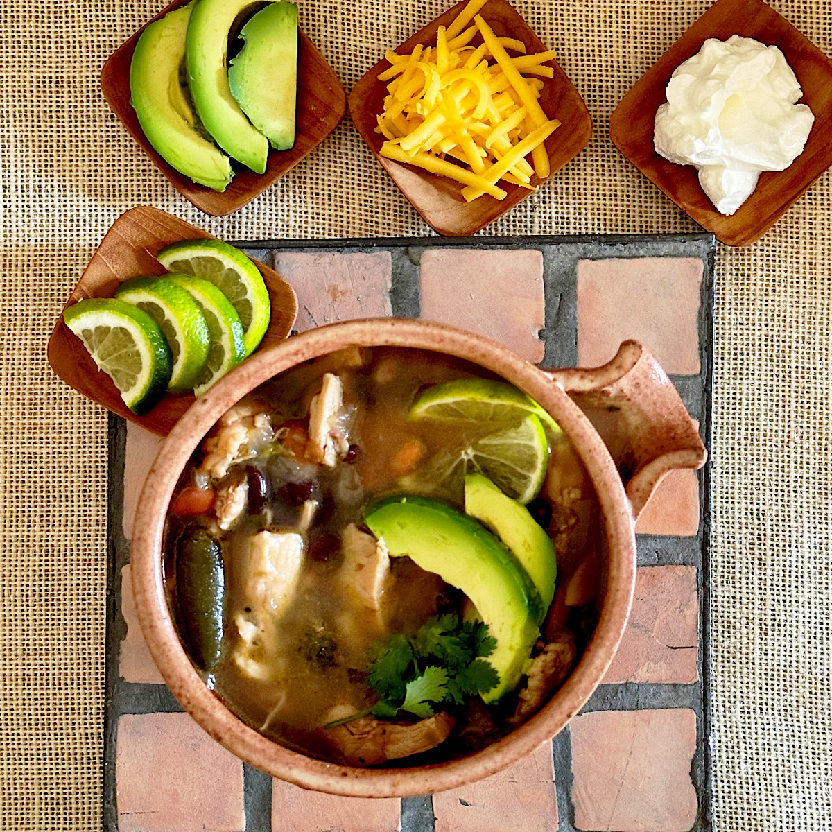 Mexican chicken chili surrounded by 4 garnishes: limes, avocados, cheese, sour cream.