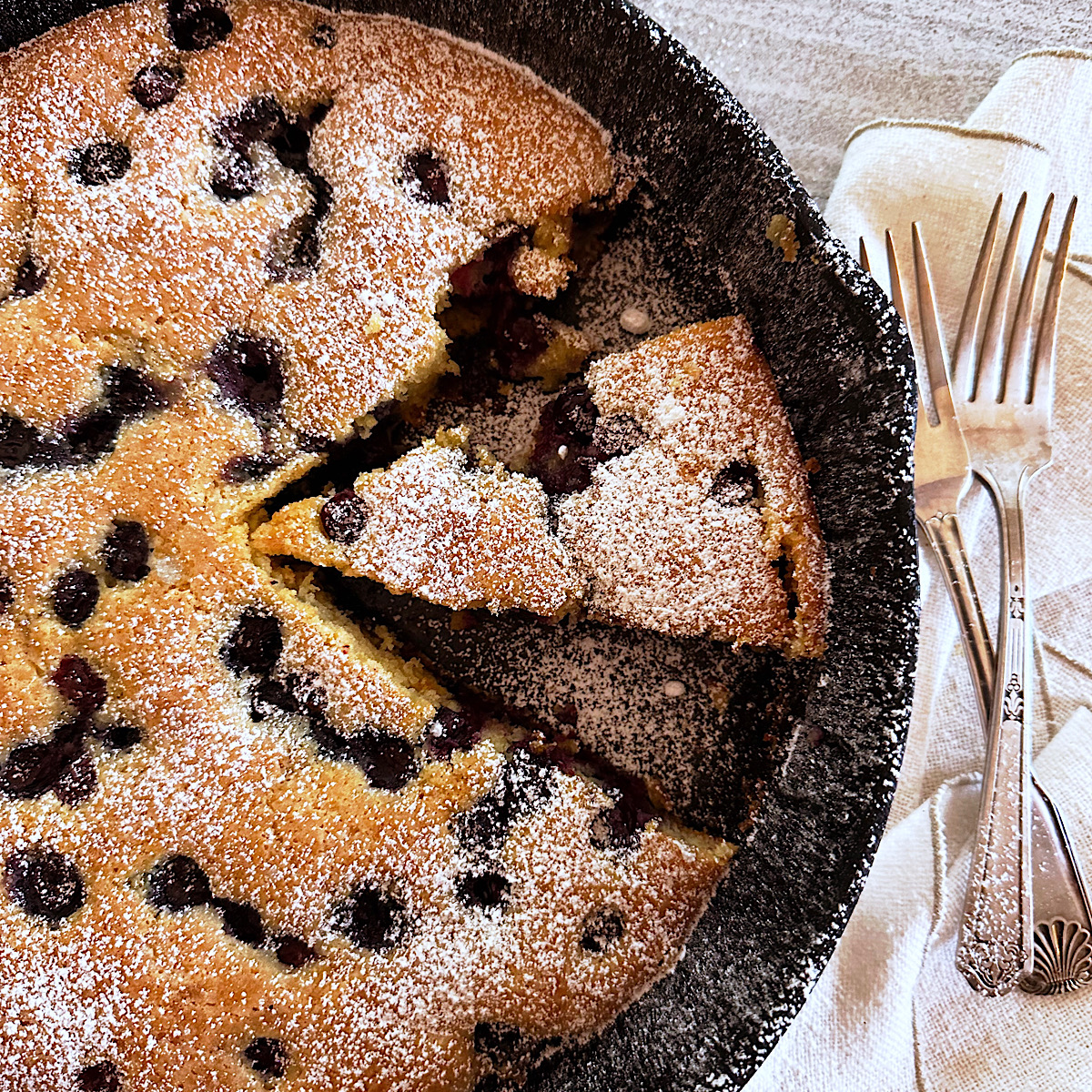 Blueberry cornmeal cake in a black skillet with a side of 2 forks.
