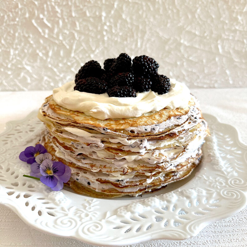 Blackberrry crepe cake on white plate with violets on side.
