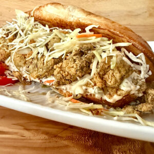 Southern Oyster Po Boy sandwich made with canned oysters, breaded in creole spice rub, deep fried and served with a caper remoulade sauce and a cabbage slaw. Served on a white plate with cabbage slaw. Served on a white plate with a side of sliced cherry tomatoes.