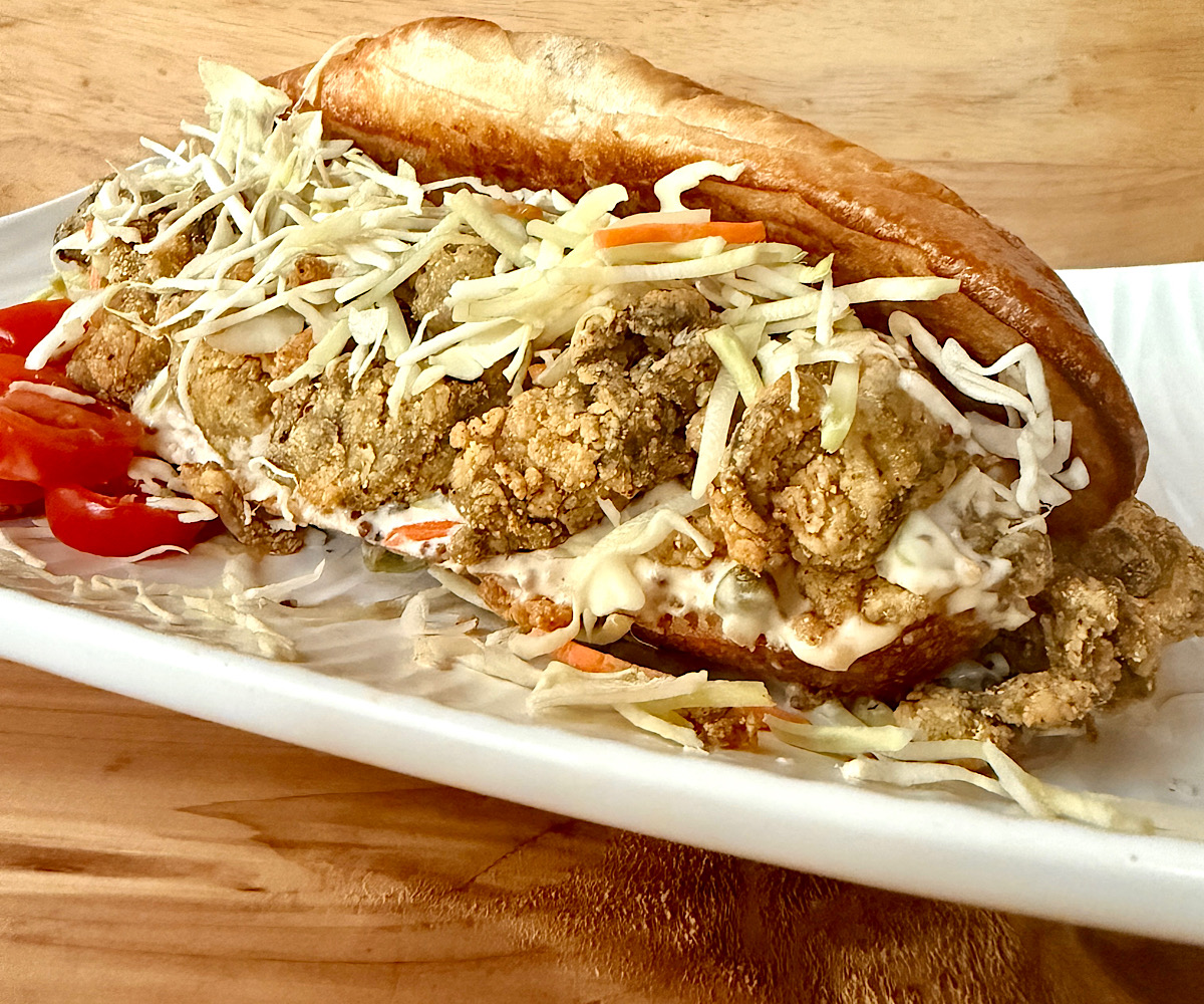 Southern Oyster Po Boy sandwich made with canned oysters, breaded in creole spice rub, deep fried and served with a caper remoulade sauce and a cabbage slaw. Served on a white plate with cabbage slaw. Served on a white plate with a side of sliced cherry tomatoes.