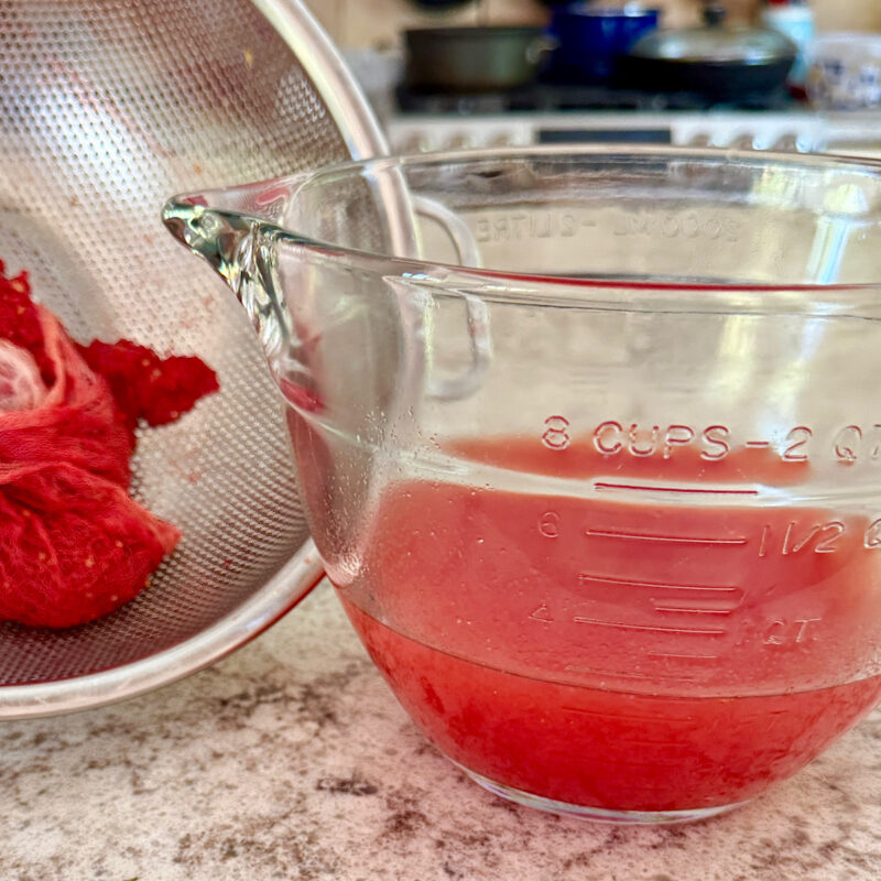 3 1/2 cups of tomato water made from fresh heirloom tomatoes from HeathGlen Farm. Colander tipped on side shows tomato pulp from straining the tomatoes through cheesecloth.