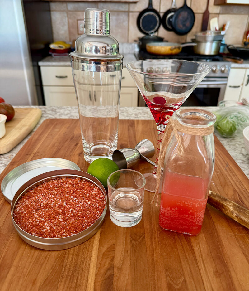 Ingredients for a tomato martini, including martini glass, tomato water, Prepar Vodka, lime for using with tomato salt for rimming, tomato water, tomato rimming salt, and shaker (photo is missing the vermouth).